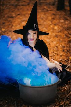 Black witch sits in front of cauldron from which smoke is falling. Sorceress conjures, brews potion with spells. Horror, halloween, cosplay holiday, magic concept. High quality photo