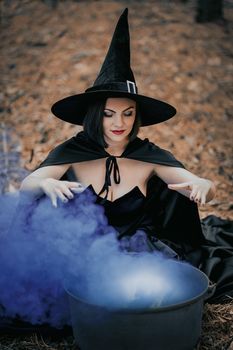 Black witch sits in front of cauldron from which smoke is falling. Sorceress conjures, brews potion with spells. Horror, halloween, cosplay holiday, magic concept. High quality photo