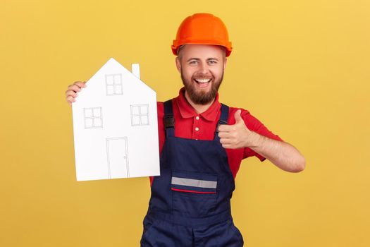 Portrait of builder man holding paper house, offering help in relocation, recommend home repair services, showing thumb up, wearing overalls and cap. Indoor studio shot isolated on yellow background.