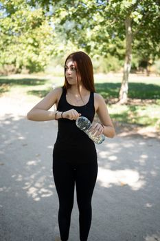 Young female athlete in black activewear opening bottle of water and looking away while standing on asphalt path during break in fitness training in park