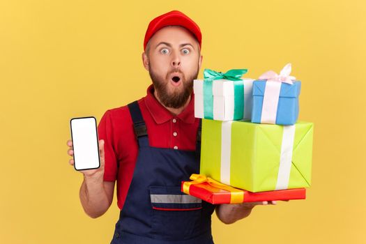 Portrait of shocked courier man wearing blue overalls holding stack of present boxes and smart phone with blank screen for advertisement. Indoor studio shot isolated on yellow background.