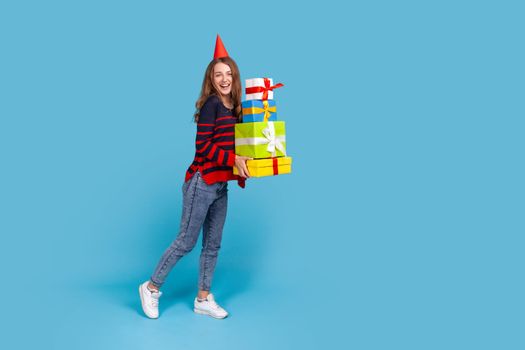Full length portrait of woman wearing striped casual style sweater and red party box standing with many present boxes, looking at camera with excitement. Indoor studio shot isolated on blue background