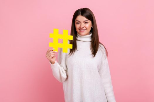 Beautiful woman holding hashtag symbol, promoting viral topic in social network, tagging blog trends, wearing white casual style sweater. Indoor studio shot isolated on pink background.