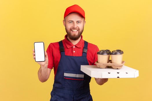 Portrait of smiling deliveryman standing with pizza box and coffee in disposable cup and showing smart phone with empty display for advertisement. Indoor studio shot isolated on yellow background