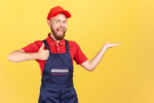 Portrait of optimistic handyman standing and presenting copy space for advertisement on his palm, showing thumb up, wearing overalls and red cap Indoor studio shot isolated on yellow background.