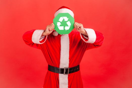 Unknown man with wearing santa claus costume and red hat, standing covering his face with green recycling sign, environment, ecology. Indoor studio shot isolated on red background.