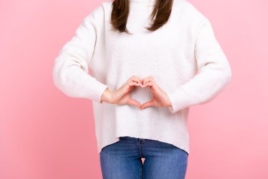 Unknown girl making heart shape in front of her belly as symbol of women health care, healthy eating, wearing white casual style sweater and jeans. Indoor studio shot isolated on pink background.