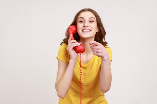 Smiling attractive woman of young age in yellow t-shirt pointing finger on you holding in hands landline telephone, waiting for your call. Indoor studio shot isolated on gray background.