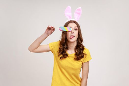 Portrait of adorable funny teenager in yellow casual T-shirt with pink bunny ears looking at camera and covering eye with ice cream. Indoor studio shot isolated on gray background