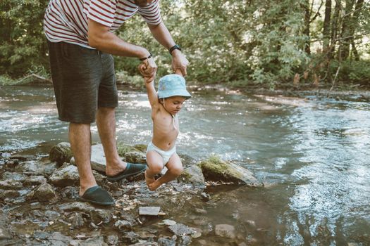 Dad holds his little baby over the river. Funny son tied his legs and does not want to swim in water. High quality photo