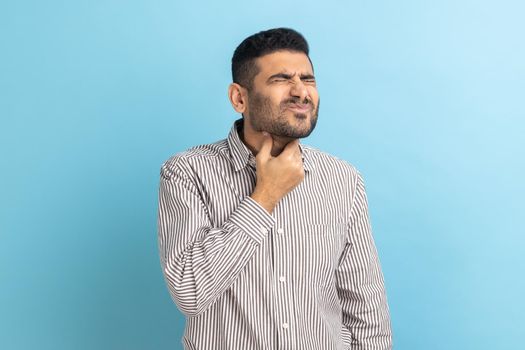 Throat pain. Portrait of unhappy ill businessman touching her neck, suffering sore throat, viral infection or flu symptoms, wearing striped shirt. Indoor studio shot isolated on blue background.