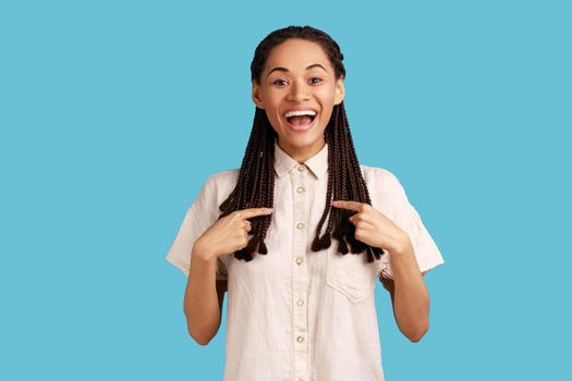Cheerful excited woman with black dreadlocks pointing at herself and smiling broadly, extremely happy being chosen, wearing white shirt. Indoor studio shot isolated on blue background.