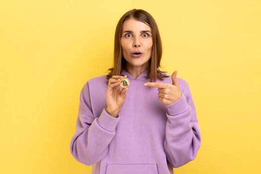 Cryptocurrency, digital money. Surprised excited woman showing golden bitcoin and looking with amazement, pointing at btc coin, wearing purple hoodie. Indoor studio shot isolated on yellow background.