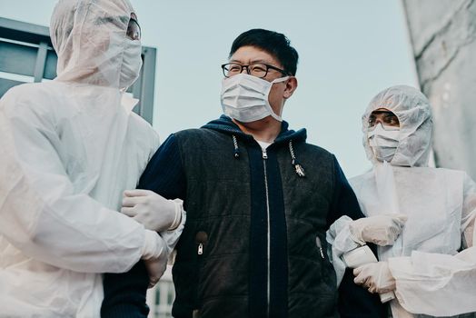 Asian man breaking covid regulation getting taken away or arrested by healthcare workers wearing hazmat protective suits. Male removed for not following the rules or restrictions of the pandemic
