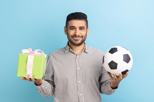 Portrait of happy delighted businessman standing looking smiling at camera, holding soccer ball and present box, wearing striped shirt. Indoor studio shot isolated on blue background.