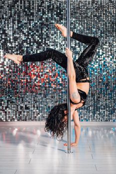 Woman doing tricks, practicing in studio with pylon. Young stripper in black dances sexually with pole in club. Poledance on shining wall background. High quality photo
