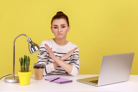 Upset sad woman manager worker sitting at workplace crossing hands showing x sign, rejecting and finishing sexual harassment at work. Indoor studio studio shot isolated on yellow background.