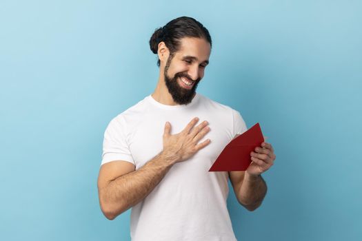 Portrait of man with beard wearing T-shirt reading letter from red envelope, gets greeting card, having positive romantic expression, being pleased, Indoor studio shot isolated on blue background.