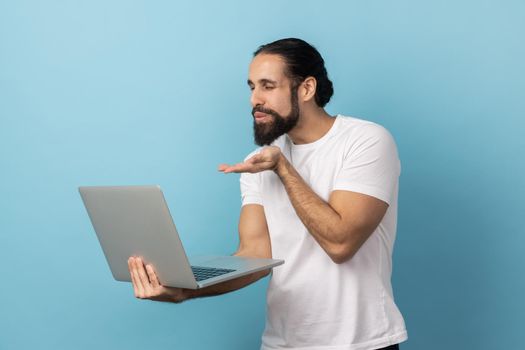 Love you. Portrait of man with beard wearing white T-shirt sending air kiss to laptop screen, talking on video call, expressing romantic feelings. Indoor studio shot isolated on blue background.