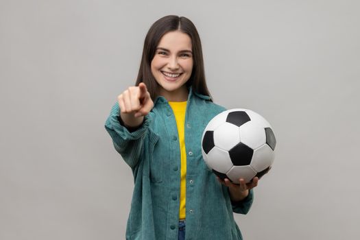 Young beautiful dark haired woman holding soccer ball, looking at camera with toothy smile, pointing finger to camera, wearing casual style jacket. Indoor studio shot isolated on gray background.