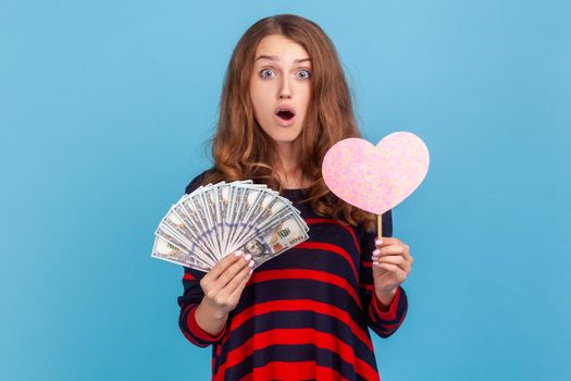 Astonished young adult woman wearing striped sweater, showing pink heart on a stick and fan of dollars, looking at camera with shocked expression. Indoor studio shot isolated on blue background.