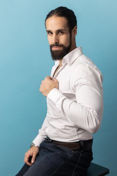 Side view portrait of strong confident man with beard wearing white shirt and black trousers, looking at camera with mysterious look. Indoor studio shot isolated on blue background.