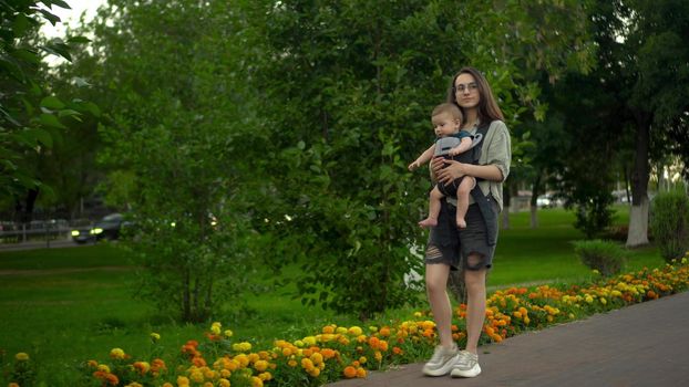 A young woman walks along the alley in the park with her son. The girl in glasses walks. Newborn son in a kangaroo-backpack. 4k