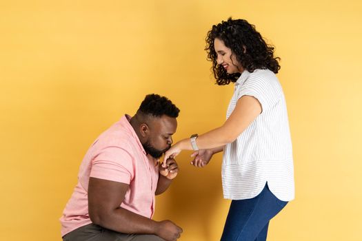 Portrait of romantic young couple in casual clothing posing together, man standing on his knee and kissing her wife hand, expressing love. Indoor studio shot isolated on yellow background.