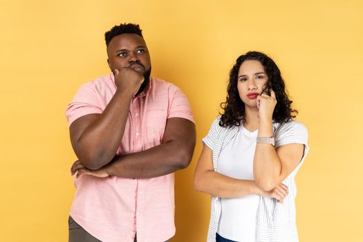Portrait of thoughtful young couple in casual clothing standing together, holding their chins while thinking solution, looking away. Indoor studio shot isolated on yellow background.