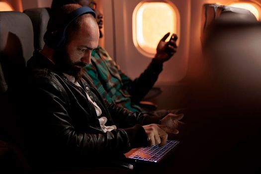 Diverse group of passengers flying in economy class on plane jet, travelling to holiday destination. Using laptop and smartphone during sunset flight before arriving on vacation trip.