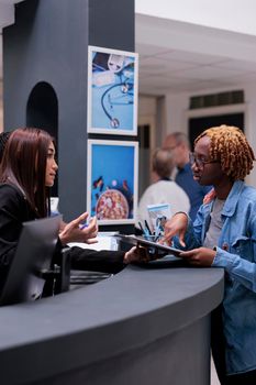 Female receptionist helping woman at reception desk, signing medical report papers to receive insurance support before checkup appointment. Patient filling in registration form to do examination.