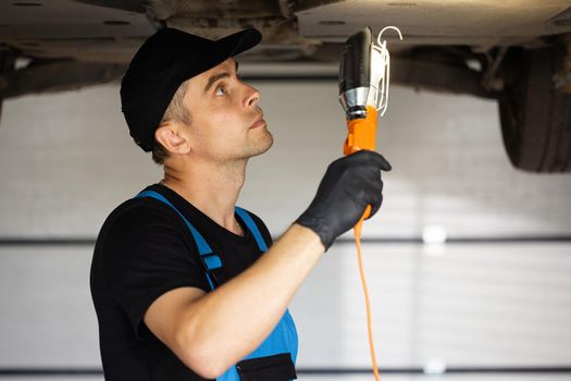 Auto mechanic man in blue overalls with lamp working at workshop. Professional mechanic is working on a car in a car service. Car service, repair, maintenance and people concept.