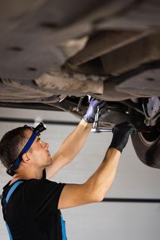 Handsome professional car mechanic is working under a vehicle on a lift in service. Repairman is using led lamp and takes a ratchet. Modern workshop.