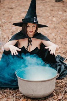 Black witch sits in front of cauldron from which smoke is falling. Sorceress conjures, brews potion with spells. Horror, halloween, cosplay holiday, magic concept. High quality photo