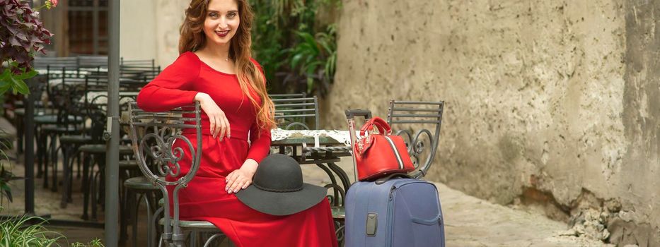 Beautiful young caucasian woman in a red long dress with hat sitting and posing looking at camera in a cafe outdoors