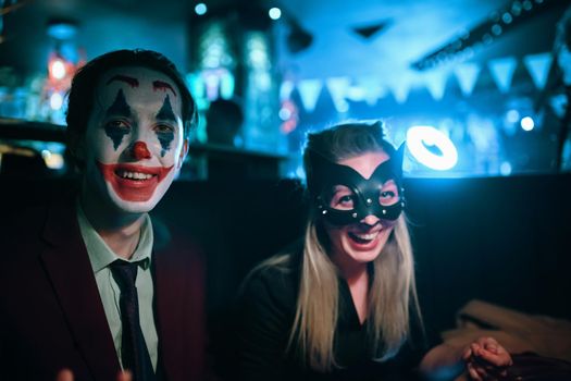 Woman in cat mask and guy with joker makeup sits at table in pub. Halloween party at a nightclub. All Saints' Night. Garland lights on the background. Photo card with copy space.