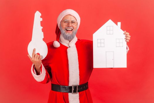Smiling elderly man with gray beard wearing santa claus costume holding in hands paper house and key, being happy to buy new apartment. Indoor studio shot isolated on red background.