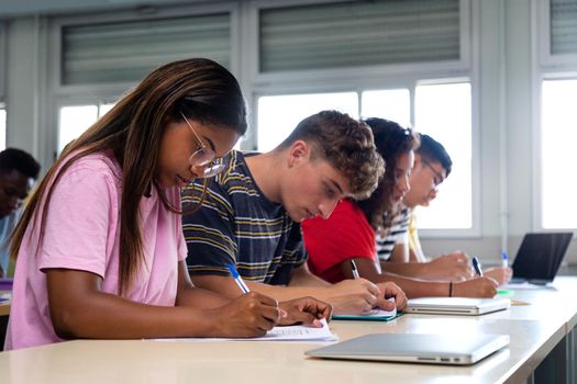 African american teen high school student writing, doing homework in class with multiracial classmates. Group of college students taking notes in class. Education.