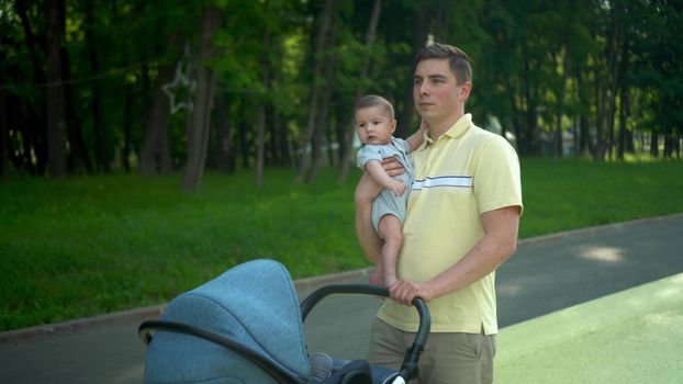 A young dad holds a newborn baby in his arms and carries a stroller. A man and his son are walking in the park against the backdrop of the forest. 4k
