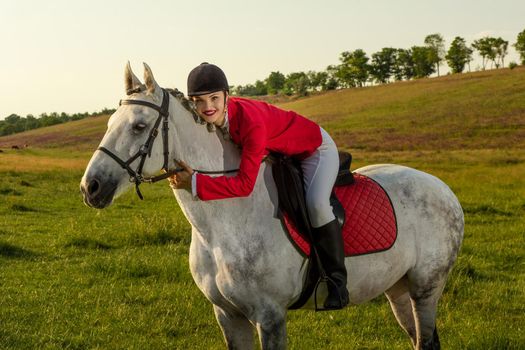 Young woman rider, wearing red redingote and white breeches, with her horse in evening sunset light. Outdoor photography in lifestyle mood