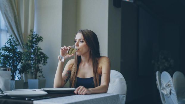 Pretty young woman is sitting alone at table in restaurant, drinking champagne and waiting for her boyfriend. Girl wearing beautiful dress is nervous and feeling uncomfortable.