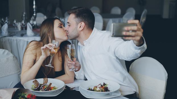 Attractive loving couple is taking selfie with champagne glasses using smartphone while having romantic dinner in restaurant. They are smiling, kissing and posing looking at camera.