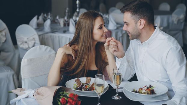 Happy loving couple is holding hands, talking and kissing during romantic dinner in luxurious restaurant. Affectionate relationship, love and fine dining concept.