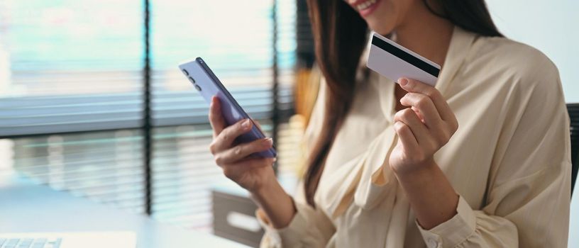 Satisfied woman holding credit card, making payment on internet via smart phone. Online shopping, e-commerce concept.