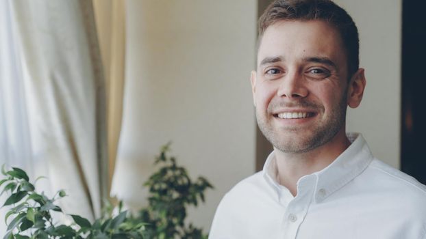 Close-up portrait of dark-haired handsome young man in white shirt smiling happily and looking at camera. Young people, beautiful interior and happiness concept.