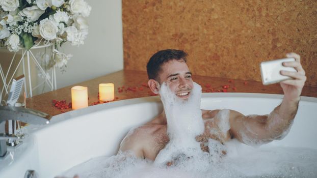 Cheerful young man with foam on beard is taking selfie using smartphone in hot tub in modern spa salon. He is laughing, gesturing, posing and looking at camera.