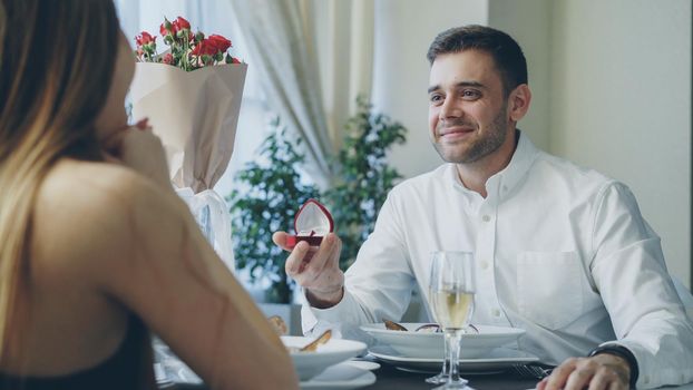 Well dressed handsome guy is making a proposal to young lady while dining in restaurant. He is talking then giving her engagement ring in jewelry box, woman is happy.
