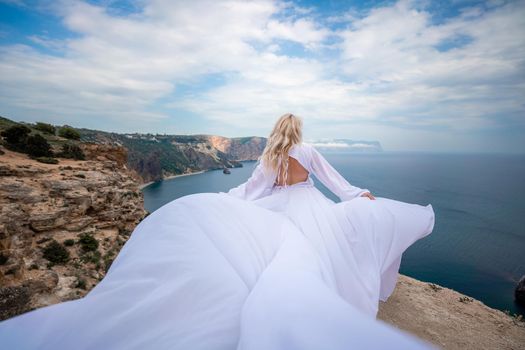 Blonde with long hair on a sunny seashore in a white flowing dress, rear view, silk fabric waving in the wind. Against the backdrop of the blue sky and mountains on the seashore