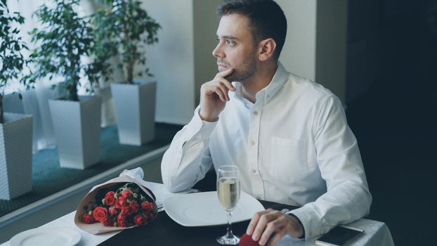 Well-dressed angry young man is waiting for his girlfriend in restaurant, bouquet of flowers left on table.