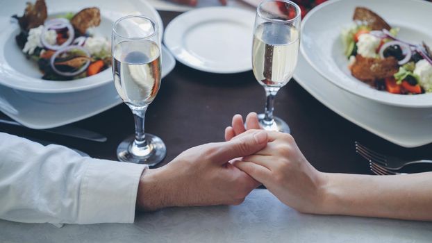Close-up shot of male hand holding and caressing manicured female hand on table with champagne glasses and plates. Romantic relationship, love and fine dining concept.